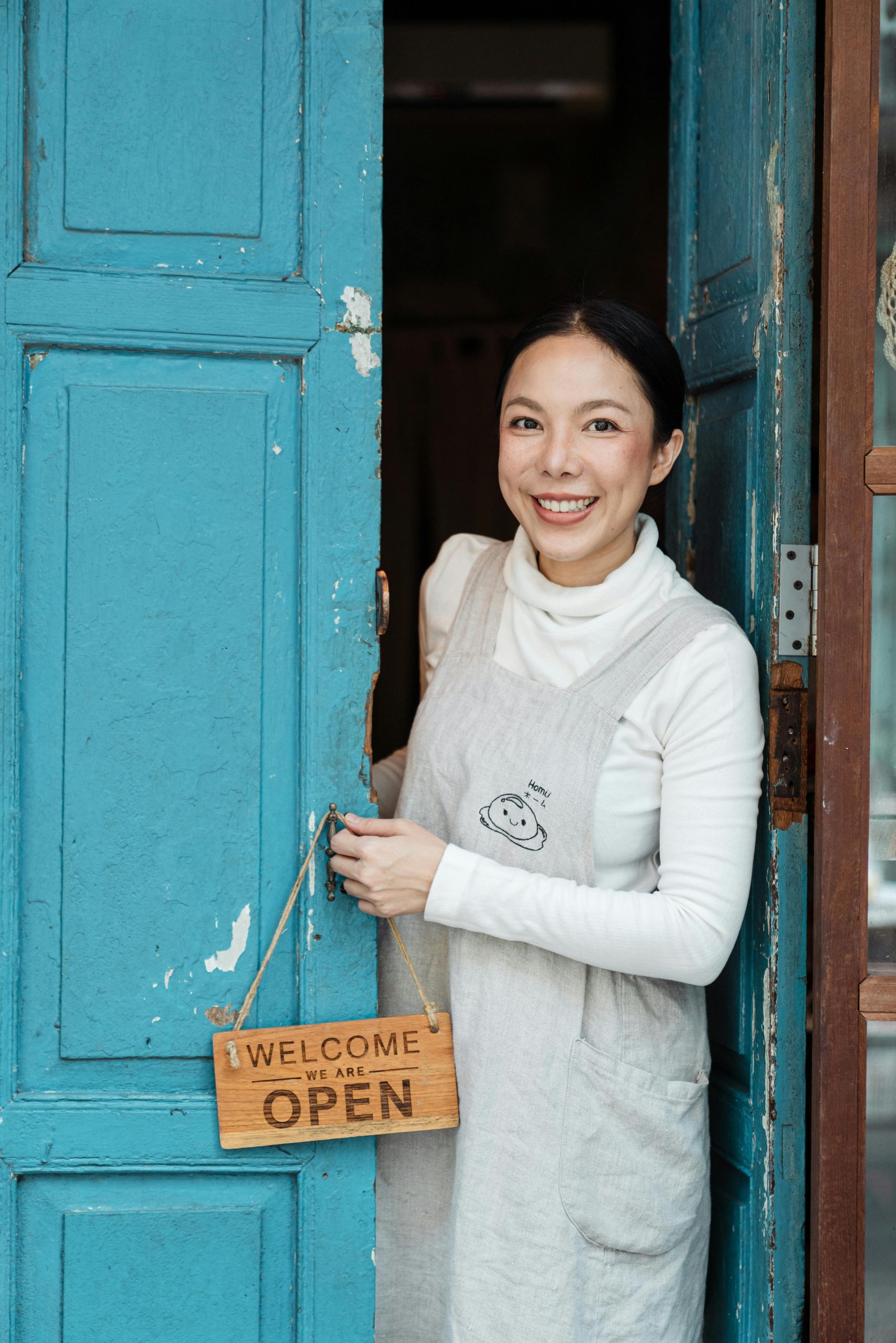 Friendly woman standing at a rustic blue door, holding a welcome sign, inviting ambiance.